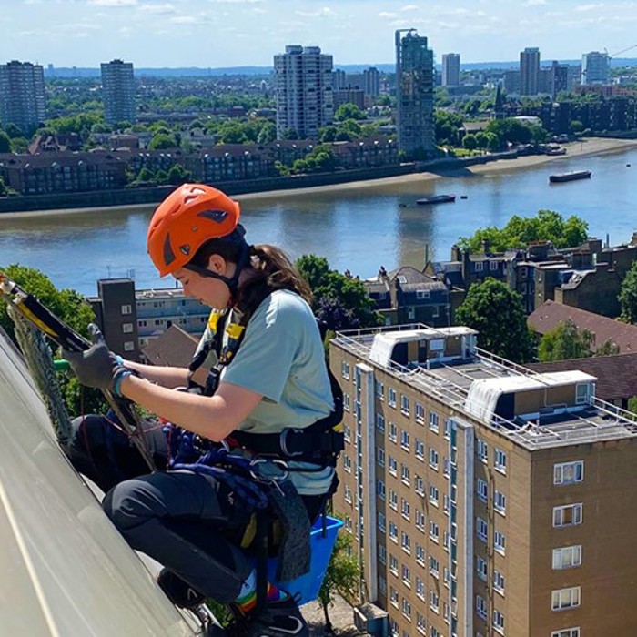 Person wearing safety gear is cleaning the windows of a tall building with a river and cityscape in the background.