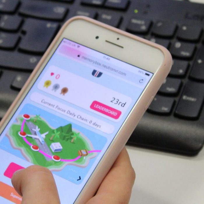 A person holds a smartphone displaying a colorful game. The background shows a blurred computer keyboard and mouse.