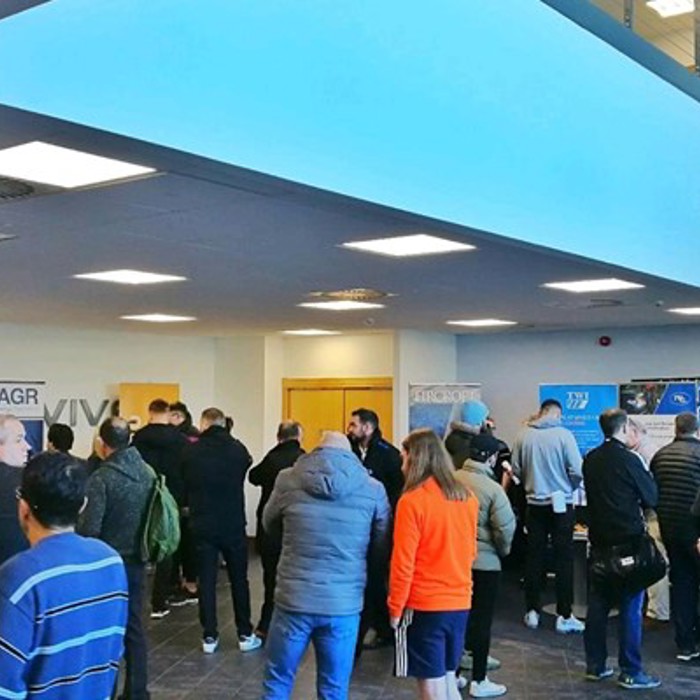A group of people, some in conversation, stand in a spacious, well-lit lobby area with various informational banners and posters displayed around.