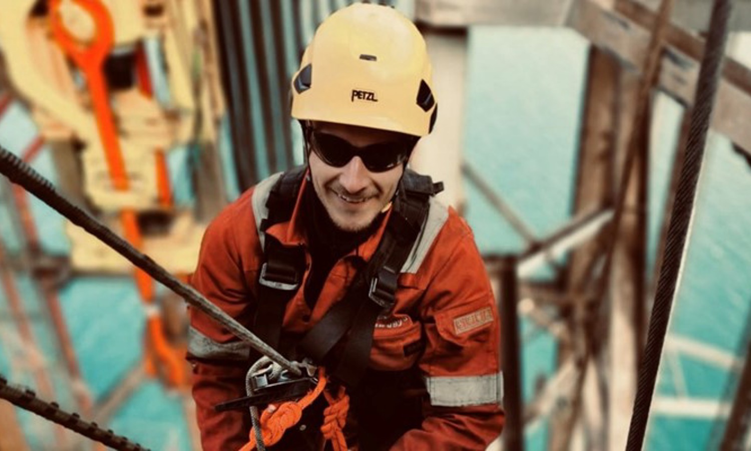 Um trabalhador com um capacete amarelo e equipamento de segurança está a subir uma estrutura com cordas, com uma grande massa de água visível ao fundo.