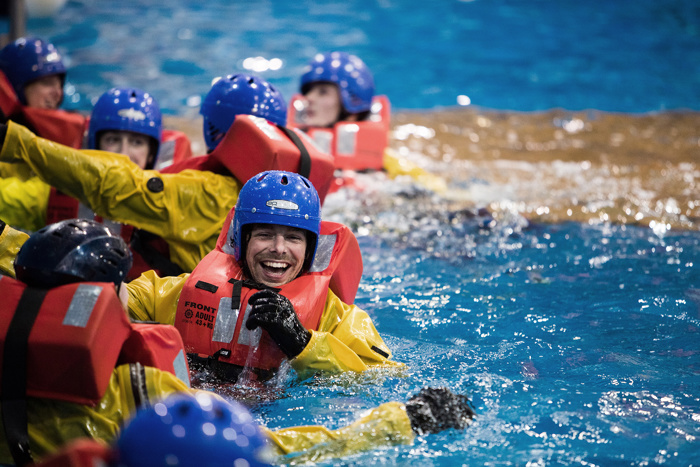As pessoas que usam coletes salva-vidas e capacetes participam numa formação sobre segurança na água, sorriem e interagem numa piscina.