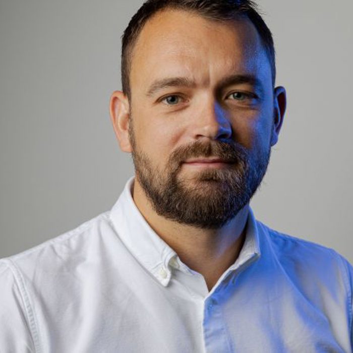 A man with a short beard and short hair wearing a white shirt stands against a plain gray background, looking at the camera.