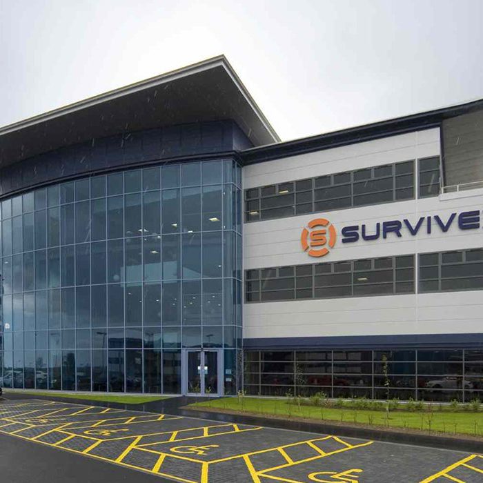 Modern commercial building with large glass facade and "Survivex" sign, featuring a parking lot with marked spaces and several parked cars in front.