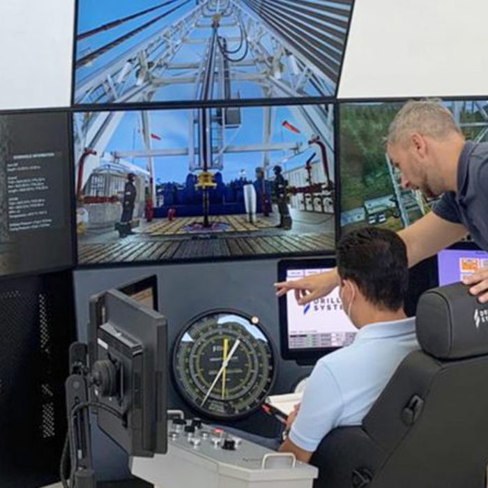 Two men in a control room monitor multiple screens displaying drilling operations. One seated man operates controls while another man stands beside him, pointing to a screen.
