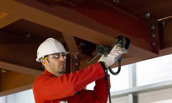 A worker in a red jumpsuit and white helmet uses a power tool on a steel beam, standing on scaffolding indoors.