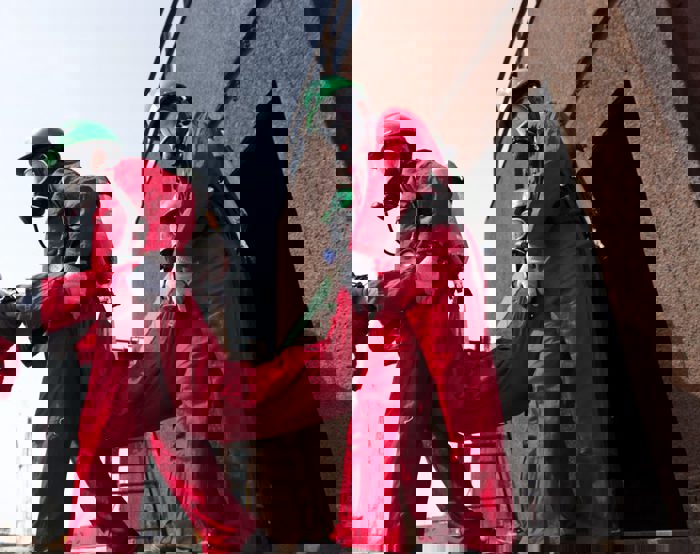 Emergency responders in red protective suits and green helmets carry an individual up a metal staircase.