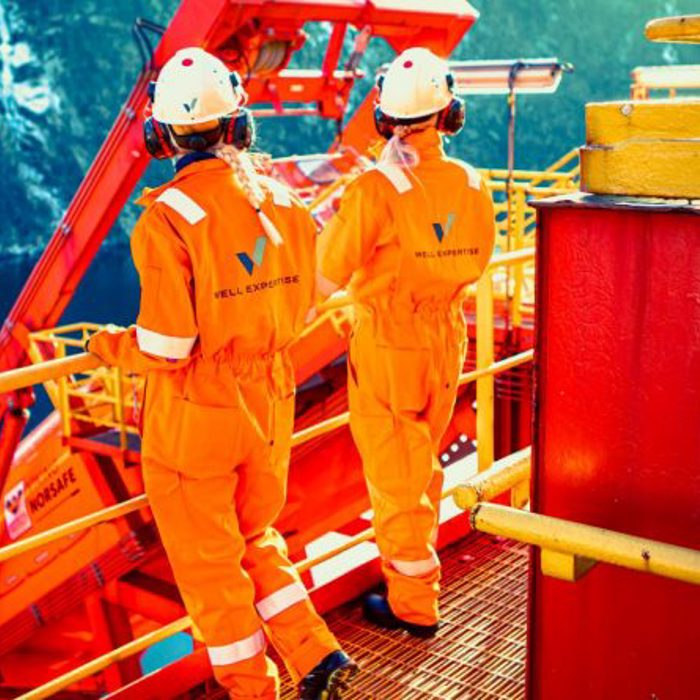 Two workers in orange safety uniforms and helmets standing on an offshore oil rig platform, facing away with equipment and safety railing visible around them.