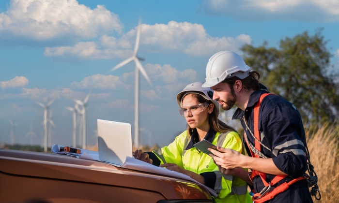 Two technicians in safety gear and helmets work on a laptop on the back of a vehicle, with wind turbines in the background under a partly cloudy sky.