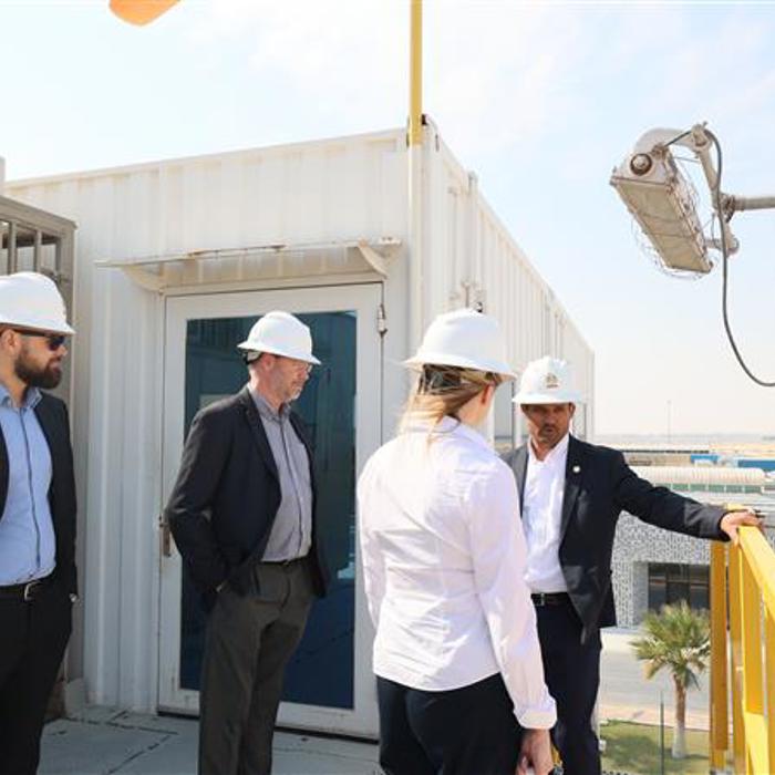 Four people in hard hats at an industrial site, standing on a platform near equipment and a building.
