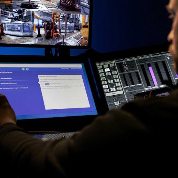 A person operates machinery using multiple computer screens and control panels in a control room, with industrial equipment visible on the monitors.